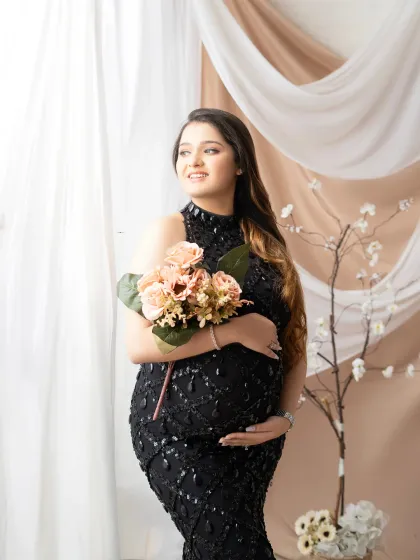 Glamour and grace. This expectant mother looks stunning in a sparkling black halter gown, holding a bouquet of flowers in a softly lit studio setting.