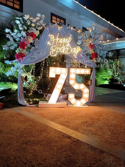 A grand celebration for a 75th birthday. This outdoor setup features large, brightly lit marquee numbers, a 'Happy Birthday' neon sign, and an arch decorated with white drapes and flowers for an elegant touch.