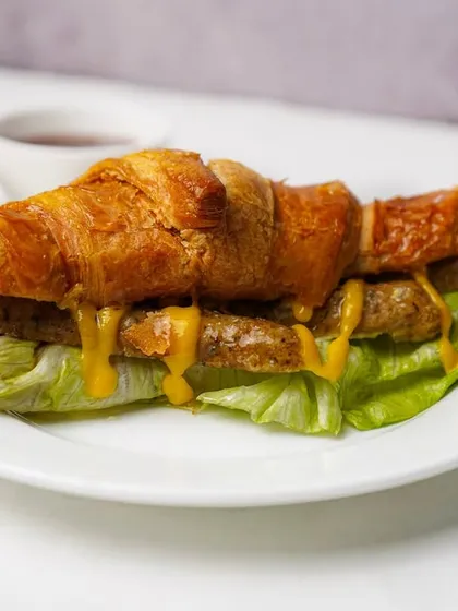 A close-up of a sausage croissant sandwich, highlighting the texture of the bread and the melted cheese. This clean shot is designed to make the item look as tasty as possible for online ordering.