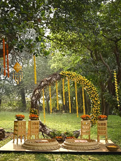 A rustic and natural Haldi setup in a wooded area. The centerpiece is a circular arch made of twigs and decorated with marigold garlands, creating a beautiful blend of rustic charm and traditional elements.