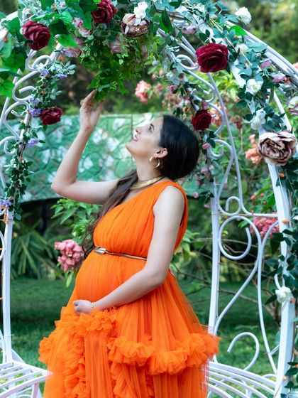A candid moment of a mom-to-be in an orange gown, reaching up to touch the flowers on a beautiful white arch. It's a lovely, interactive shot.