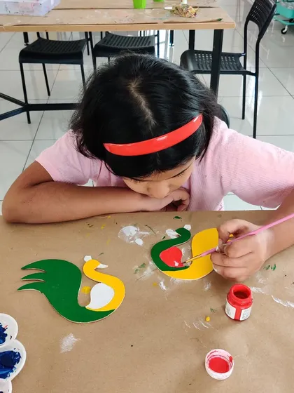 Close-up of a child painting peacock T-light holders, adding red details to the bright yellow and green design.