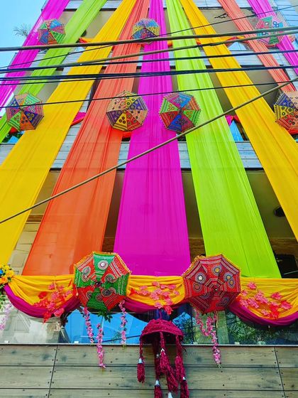 A stunning display of multi-coloured drapes and Rajasthani umbrellas adorning the front of a building for a wedding celebration.