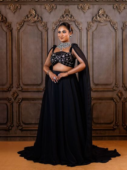 A full-length view of a modern black lehenga with a cape. The ornate wall backdrop adds a touch of classic architecture to this contemporary fashion look.