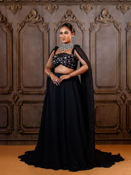 A full-length view of a modern black lehenga with a cape. The ornate wall backdrop adds a touch of classic architecture to this contemporary fashion look.