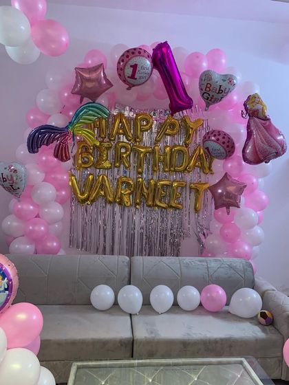 A princess-themed first birthday decoration for a home celebration. The setup includes a pink and white balloon wall, 'Happy Birthday' foil letters, and various character balloons like a mermaid tail and a princess.