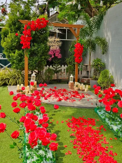 A charming garden proposal featuring a rustic wooden arch decorated with red flowers. The ground is covered in red petals, leading to the spot where a "Will you Marry Me?" sign hangs, creating a sweet and natural setting.