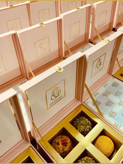 Personalized wedding favor boxes with the couple's monogram, opened to reveal a selection of delicious Indian sweets. The pastel pink and gold design adds a touch of modern romance.