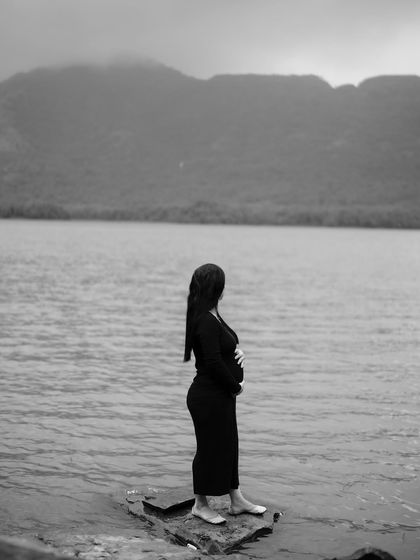 A serene black and white shot of the mom-to-be looking out over the water, a moment of quiet contemplation in a vast, beautiful landscape.