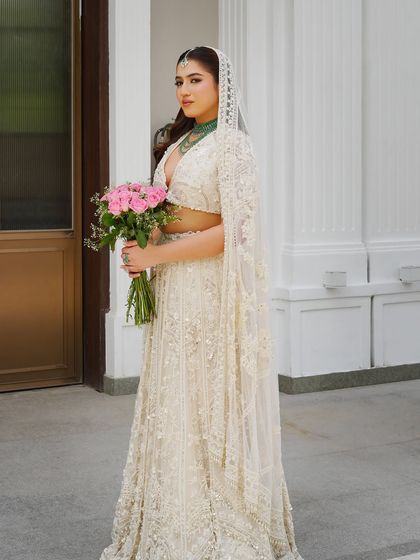 Ivory dreams and emerald accents. This eclectic and elegant look features a bride in a stunning white lehenga, holding a simple bouquet of pink roses that adds a soft pop of color.