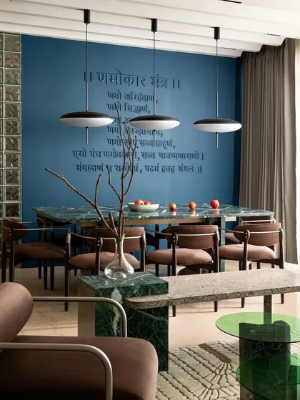 The dining area, where the green marble table and chairs are set against the serene blue wall with the Jain mantra. The pendant lights add a modern touch to the space.