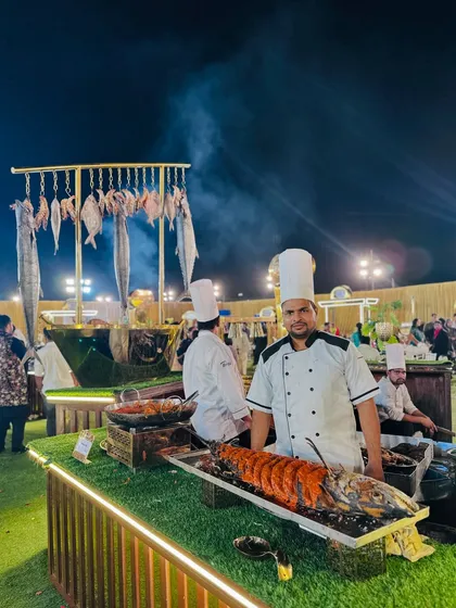 Our impressive live seafood station, where guests can see the fresh catch of the day. Here, a chef proudly presents a large grilled fish, with a backdrop of hanging fish and the sizzle of the grill, promising a fresh coastal feast.