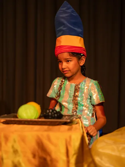 A young performer in a colorful costume, looking on with a thoughtful expression. Listening and reacting are as important as speaking in theatre.
