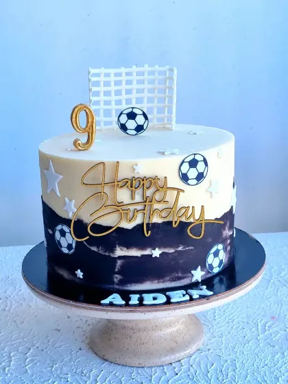 A stylish football cake with a fault line design in black and white. The cake is topped with a gold "Happy Birthday" sign and a fondant soccer goal and ball.