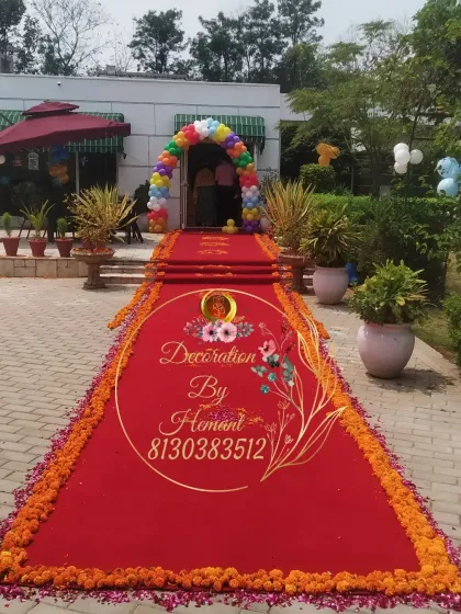 A grand entrance for a baby boy's welcome party. A long red carpet, lined with marigold flowers, leads to a colorful rainbow balloon arch at the doorway, creating a spectacular welcome.