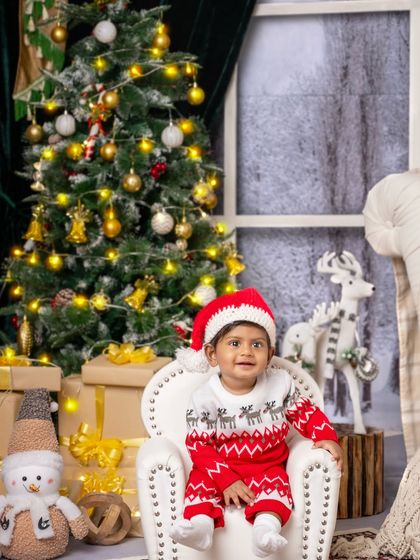 A sweet baby boy in a reindeer sweater, looking curious and happy in his mini armchair by the Christmas tree.