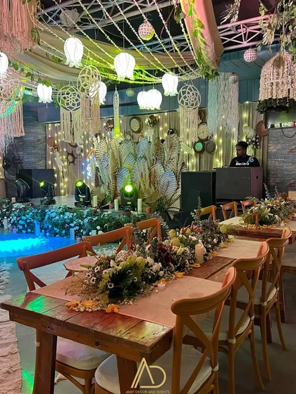 An indoor boho-themed dinner setup, with long wooden tables, rustic chairs, and an overhead installation of macrame, lanterns, and greenery.