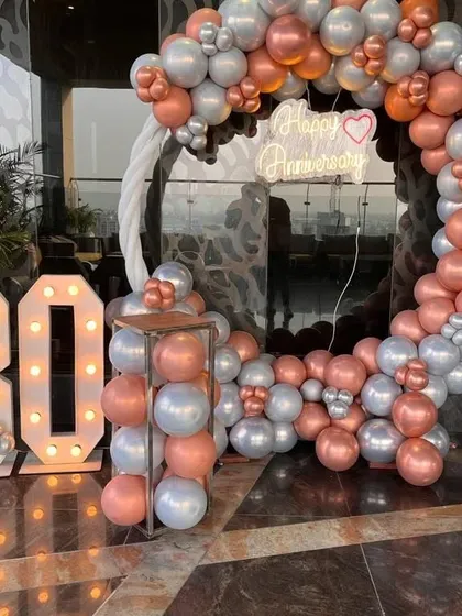 Celebrating a 30th anniversary with a stunning balloon arch against a city skyline. This setup includes a large circular arch in rose gold and silver chrome balloons, a custom 'Happy Anniversary' neon sign, and large light-up marquee numbers for the milestone year.