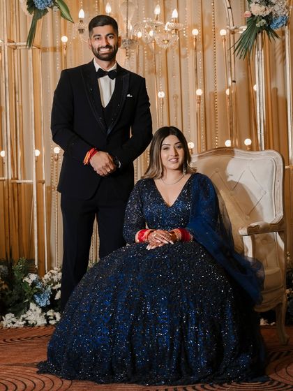 A regal portrait of the couple at their reception. The bride is seated in an elegant chair while the groom stands beside her, both dressed in stunning blue and black formalwear.