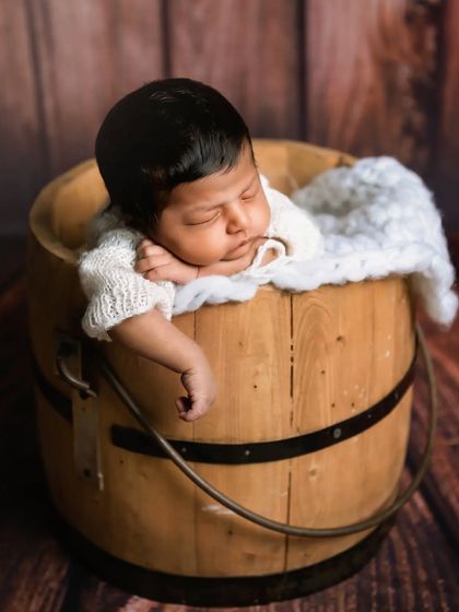 A sleeping newborn rests their head on their hands while posed in a wooden bucket. This classic prop pose is a favorite for showing how tiny the baby is.
