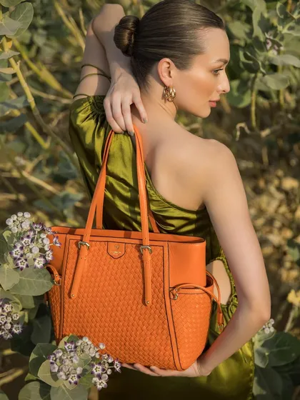A vibrant shot from the Da Milano desert campaign. The model is styled in a one-shoulder olive green dress, with the bright orange woven tote bag creating a stunning color contrast against the natural foliage.
