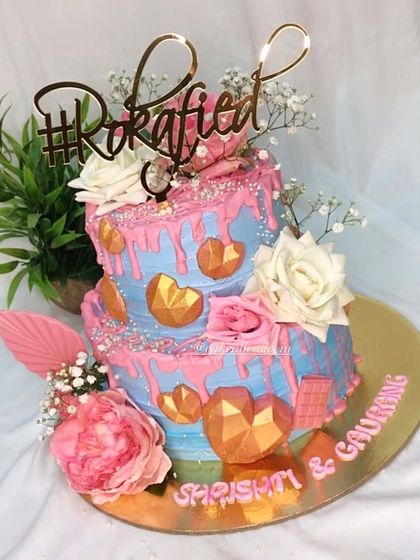 A fun and vibrant Roka cake. This two-tier design features a blue and pink color scheme, playful pink drips, and geometric chocolate hearts for a modern and celebratory feel.