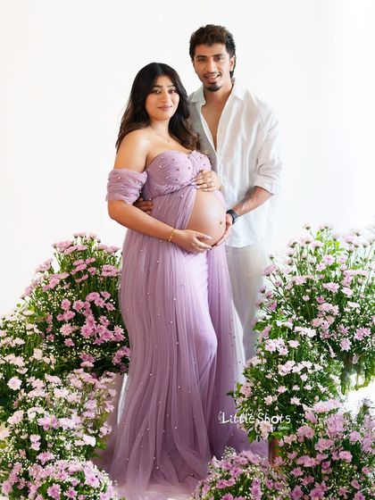 A beautiful couple's portrait set amidst flowers. The expecting mother in a lavender gown and her partner stand together, surrounded by arrangements of pink and purple flowers.