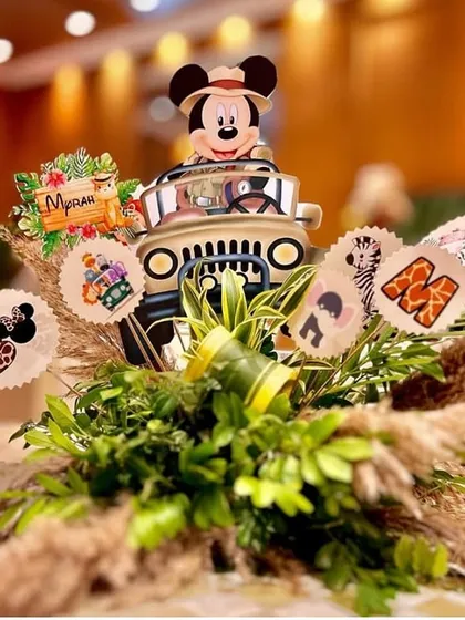 A creative jungle-themed table centerpiece. A miniature safari jeep with Mickey Mouse at the wheel is surrounded by lush greenery, pampas grass, and themed cutouts.