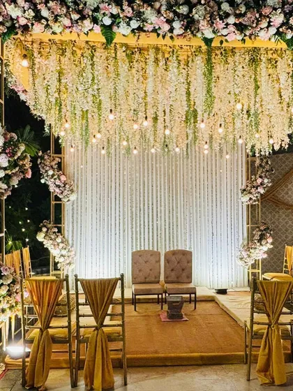 A gorgeous stage mandap with a ceiling of hanging white flowers and warm Edison bulbs. This setup is ideal for an elegant indoor or evening ceremony.
