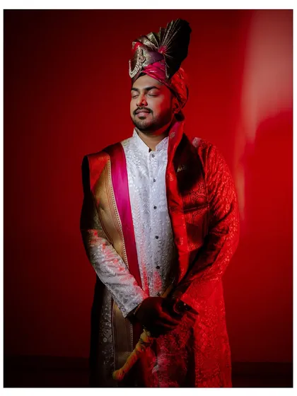 A portrait of the groom in his traditional wedding attire, captured in a moment of quiet contemplation. The red lighting adds a touch of drama and royalty.