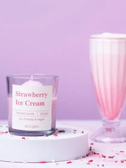 A shot of a Strawberry Ice Cream candle next to a tall milkshake glass. The composition and color coordination effectively tie the product to its scent.