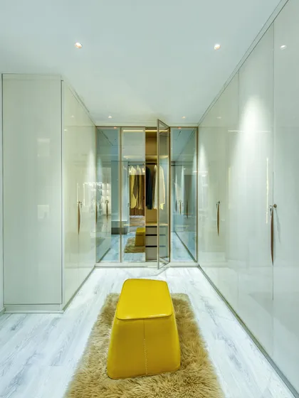 A spacious walk-in closet with sleek, handleless wardrobes and a central yellow ottoman on a plush rug. The glass-doored section allows for curated display.