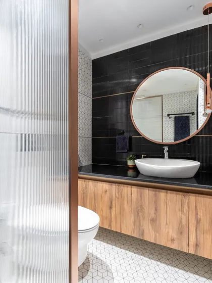 This bathroom features a modern mix of materials, including black kit-kat tiles, wood-finish vanity, and geometric floor tiles. The rose gold fixtures and round mirror add a touch of warmth and elegance.