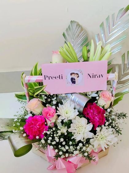 A beautiful floral arrangement curated for a couple, featuring pink roses, white chrysanthemums, and lilies in a handled basket. The personalized card and decorative leaves add a touch of modern elegance.