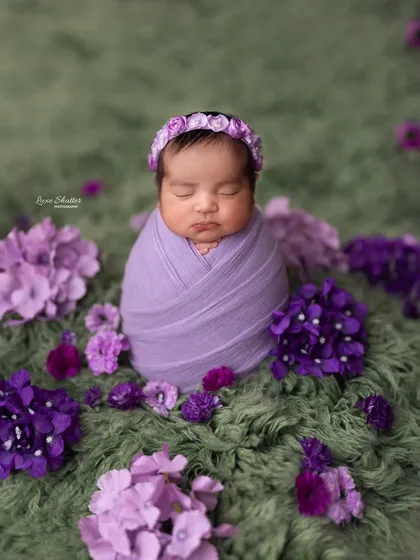 A sweet newborn swaddled in purple, nestled among a bed of flowers on a soft green rug.