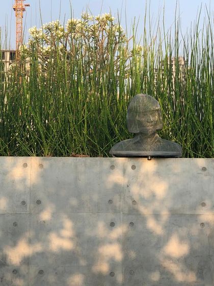 A close-up of the metal mesh bust, with dappled light filtering through the trees onto the concrete wall behind it. The image captures a quiet, contemplative mood.
