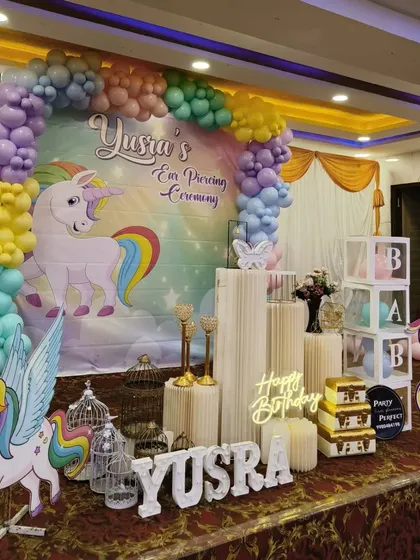 A magical unicorn-themed ear piercing ceremony and birthday party. This vibrant setup includes a large unicorn backdrop, pastel rainbow balloon arch, character cutouts, and a 'Happy Birthday' neon sign, creating a dream-like atmosphere.