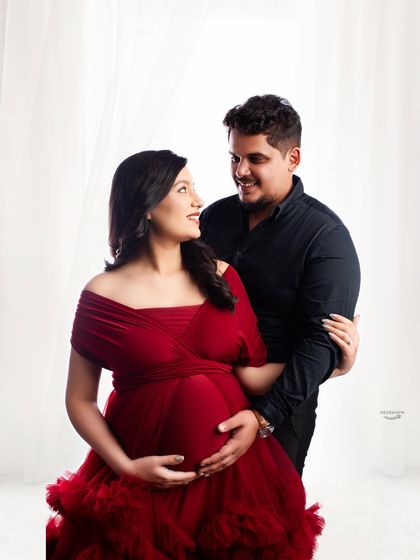 A classic and loving couple portrait. The mom-to-be is radiant in a red gown while her partner stands close, creating a timeless image of togetherness against a bright background.