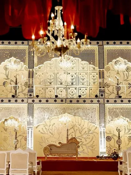 A magnificent, multi-tiered stage for a royal Jaipur reception, featuring intricate white and gold jaali work that resembles a palace facade, lit by elegant chandeliers.