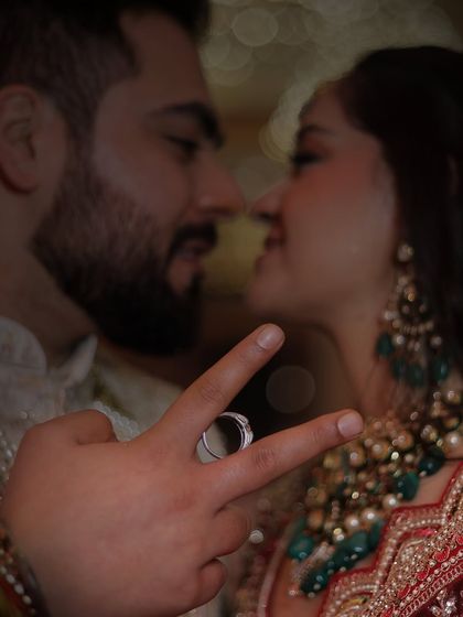 A creative and intimate couple portrait. The focus is on the engagement ring held between the groom's fingers, with the couple sharing a close moment in the background. A very artistic shot.