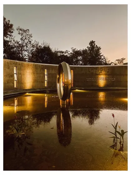 As dusk settles, the Dr. Vishnuvardhan Memorial Complex transforms. The central water body reflects the warm illumination on the concrete walls and the metallic ring sculpture, creating a serene atmosphere for evening contemplation. This is a space designed to honor memory through tranquility.