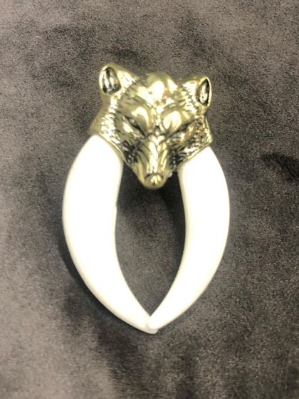 This powerful brooch features a gold-tone sculpted wolf head holding two polished white tusks. It's a distinctive accessory that adds a fierce and edgy element to any jacket or coat.