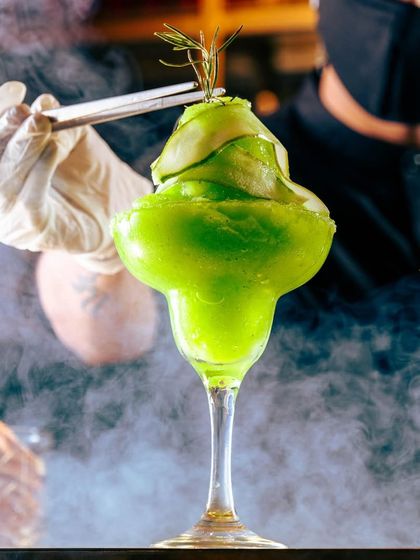 Sipping into Spring. This vibrant green frozen cocktail, garnished with cucumber and rosemary, is a refreshing ode to the awakening of nature’s palette, presented with a dramatic smoky effect.