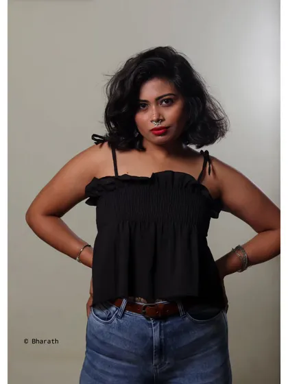 A simple and confident studio portrait of a model in a black ruffled top and blue jeans. The plain background keeps the focus on her direct gaze.