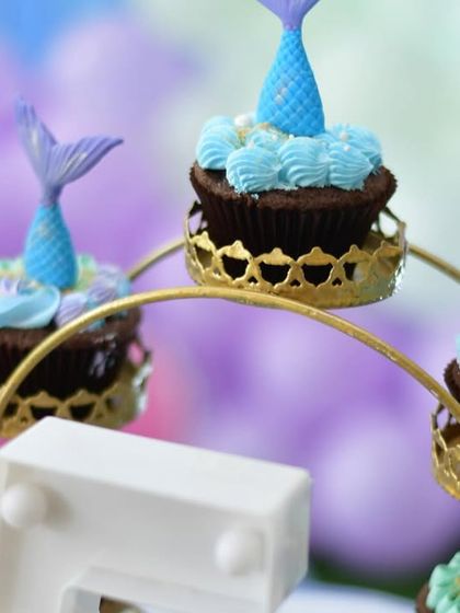 Themed cupcakes that perfectly match the mermaid party decor. Each cupcake is topped with a detailed, edible mermaid tail, displayed on a decorative ferris wheel stand.