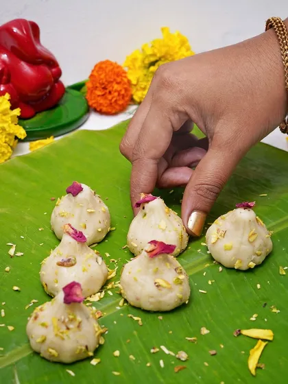 A hand arranging traditional ukadiche modak, garnished with pistachio and rose petals, on a banana leaf. This image evokes a sense of tradition and authenticity, appealing to customers looking for classic festive sweets.