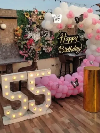 A beautiful terrace setup for a 55th birthday. This decoration features a pink and white balloon arch against a green wall, a "Happy Birthday" sign, and large marquee numbers for the age.