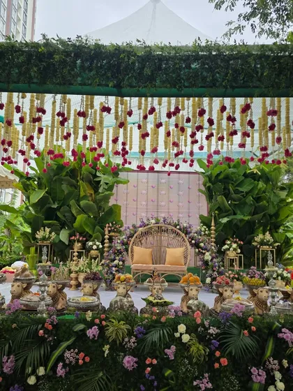 An outdoor baby shower setup with a canopy of tuberose and marigold garlands. The stage features a wicker chair and a foreground of lush flowers and traditional cow props.