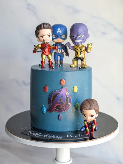 An Avengers-themed cake for a Marvel fan. The cake is a cool blue, decorated with the Avengers logo and infinity stones, and topped with fun character figurines of Iron Man, Captain America, and Thanos.
