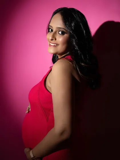 A lovely close-up portrait where the mom-to-be looks over her shoulder with a bright smile. The dramatic lighting creates a beautiful contrast, making her glow against the pink background.